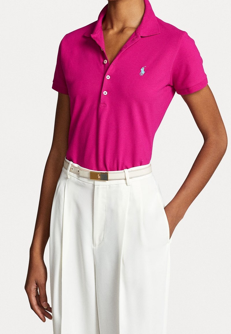 Personne portant un polo rose vif à manches courtes rentré dans un pantalon blanc taille haute avec une ceinture blanche.