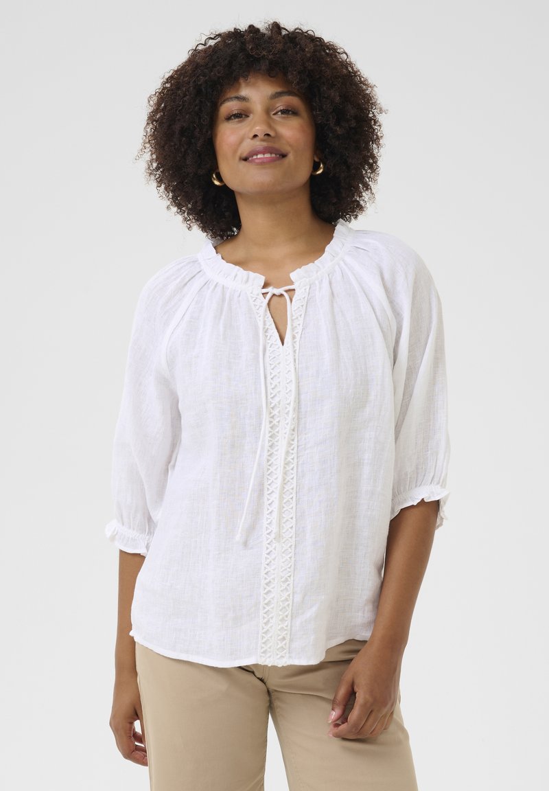 Witte linnen blouse met driekwart mouwen, geplooide halslijn en decoratieve kantafwerking langs de voorkant. Zachte textuur, losse pasvorm.