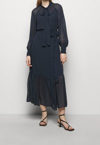 Námořnické šaty s puntíky, dlouhými průhlednými rukávy, vázáním u krku a vrstveným volánkovým lemem. Na nohou jsou černé sandály na vysokém podpatku.