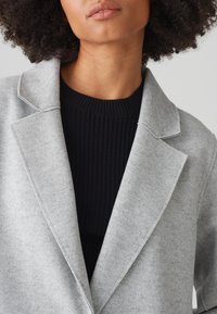 Grijze wollen blazer van een wolmix met een geknipt revers, gedetailleerde stiksels en een zachte textuur, gelaagd over een zwart geribbelde trui.