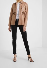 Camisa de cuero marrón con detalles de flecos y tachuelas plateadas, combinada con jeans ajustados negros y tacones altos negros con tiras.