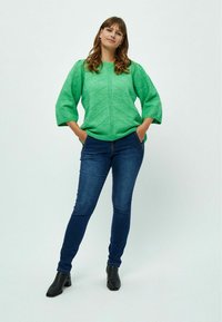 Grüner gestrickter Pullover mit strukturiertem Zickzackmuster, übergroßen Ärmeln, kombiniert mit dunkelblauen Skinny-Jeans und schwarzen Knöchelstiefeln.