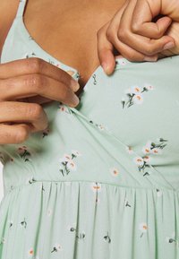 Grön blomsterklänning med vita prästkragar och orange detaljer, med en figursydd överdel, plisserad kjol och justerbart tryckknapplås.