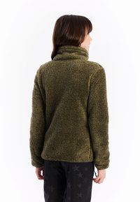 Žena otočená zády, oblečená v olivově zelené chlupaté bundě a černých kalhotách s tmavými hvězdami na bílém pozadí.