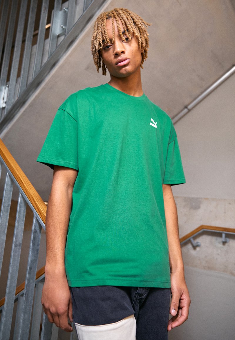 Grøn bomulds T-shirt med rund halsudskæring, korte ærmer og et lille hvidt logo på venstre bryst. Stoffet virker glat og let.