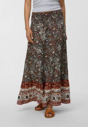 Femme portant une longue jupe à motifs floraux et cachemire dans des tons marron, blanc et rouille, associée à des sandales couleur cuir clair.