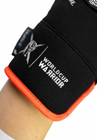 Black textile wrist guard with orange accents, featuring "WORLD CUP WARRIOR" branding and a graphic logo. Smooth texture and flexible design.