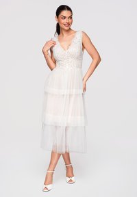 Femme souriante aux cheveux foncés, en robe blanche en dentelle sans manches et talons blancs ouverts à l'avant, debout devant un fond uni.