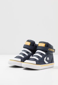 Wysokie trampki z syntetycznej skóry w kolorze granatowym, z białymi sznurowadłami i gumowymi podeszwami. Posiadają żółte akcenty oraz biały logo gwiazdy po boku.