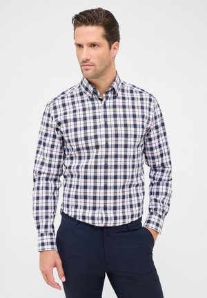 Man in een donkerblauw en wit geblokt overhemden met knopen en een donkerblauwe broek, staand tegen een lichte effen achtergrond.