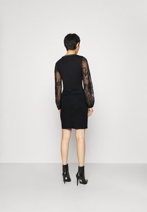 Woman with short hair wearing a black dress with sheer embroidered long sleeves and black high-heeled ankle boots, standing facing a white wall.