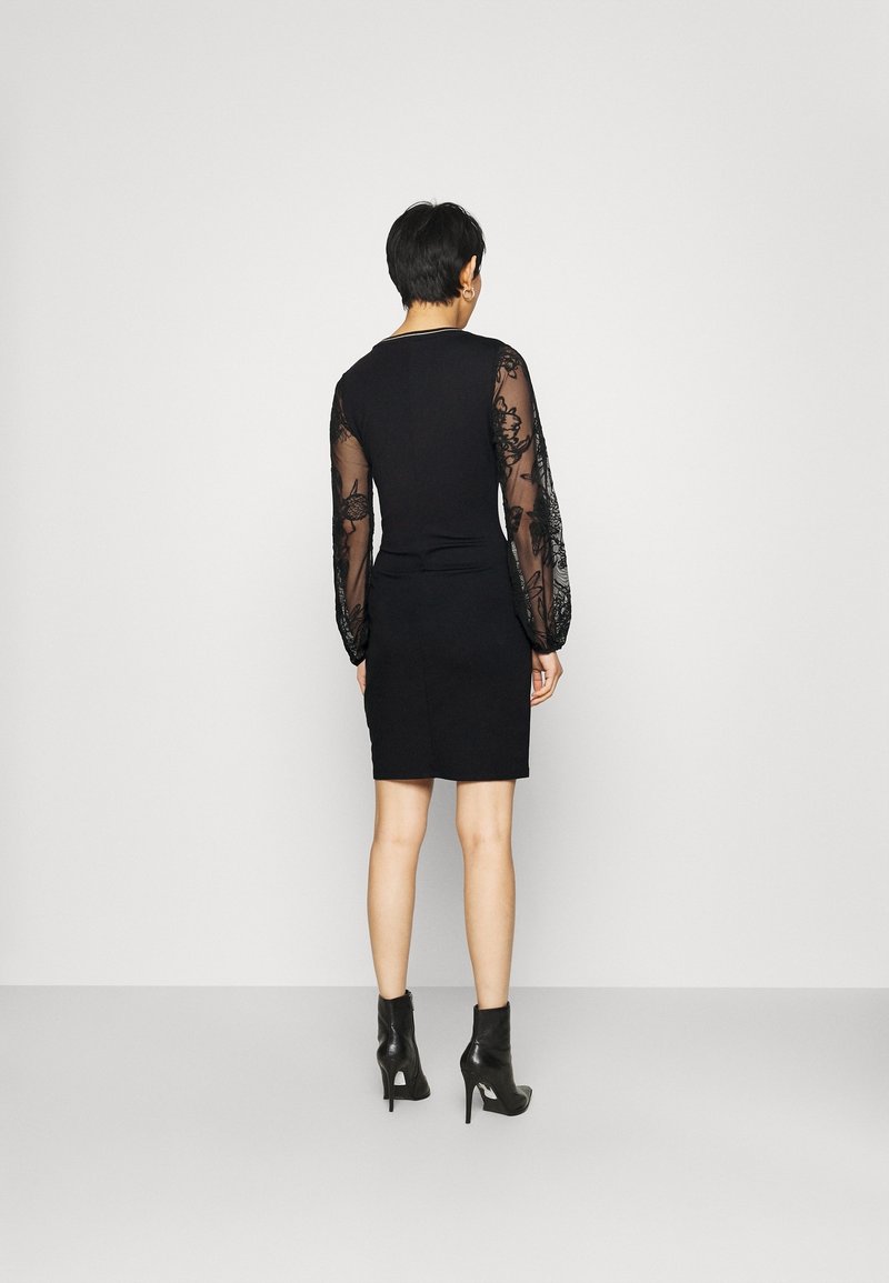 Femme aux cheveux courts portant une robe noire avec des manches longues transparentes brodées et des bottines à talons noirs, debout face à un mur blanc.