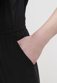 Main insérée dans la poche latérale d’un vêtement noir avec taille élastique sur un fond clair uni.