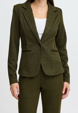 Woman wearing olive green herringbone blazer and matching trousers with a white top underneath, standing against a plain background.