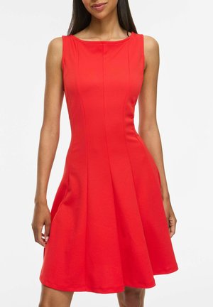 Donna con un vestito rosso senza maniche e una gonna svasata, in piedi davanti a uno sfondo bianco uniforme.