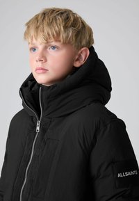 Černá péřová bunda s vysokým límcem, zipem vpředu a suchým zipem s nápisem "ALLSAINTS" na rukávu. Hladký, matný povrch s moderním designem.