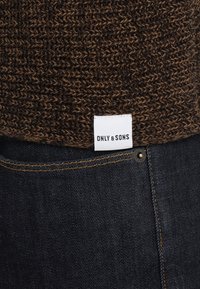 Pull en maille marron avec une étiquette en tissu blanc portant l'inscription « ONLY & SONS » au-dessus d'un jean en denim bleu foncé avec des coutures orange.