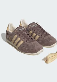 Paar braune Wildleder Adidas Japan Sneaker mit beigen Streifen, beigem Fersendetail und extra beigen Schnürsenkeln auf weißem Hintergrund.