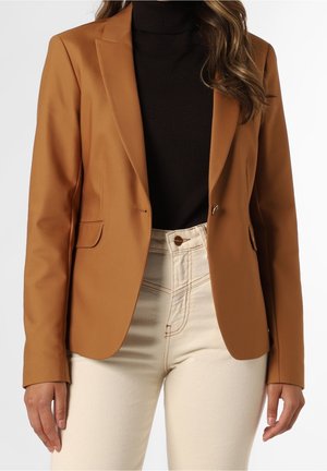 Kvinde iført en kamelfarvet blazer over en sort rullekravetrøje og højtaljede off-white bukser, der er klippet til midt på låret.