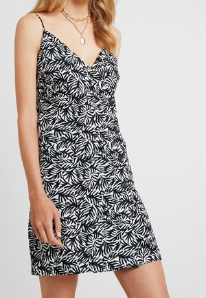 Kobieta w czarno-białej sukience na ramiączkach w liściasty wzór z guzikami po boku oraz z naszyjnikami z warstwowych złotych łańcuszków.