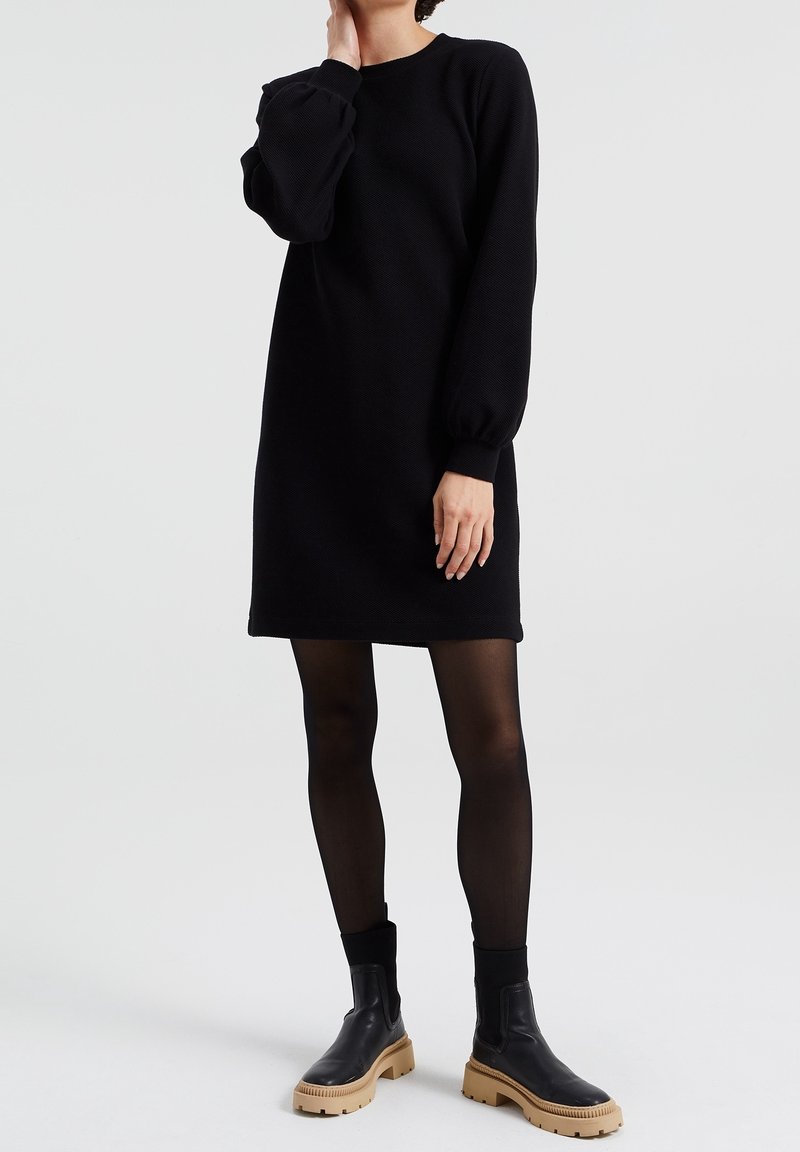 Femme en robe noire à manches longues, collants noirs et bottines noires à semelles beige, debout devant un fond clair uni.