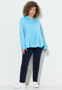 Blusa azzurra chiara a maniche lunghe con colletto e piega frontale, abbinata a pantaloni blu navy e sneakers bianche. Tessuto morbido e vestibilità rilassata.