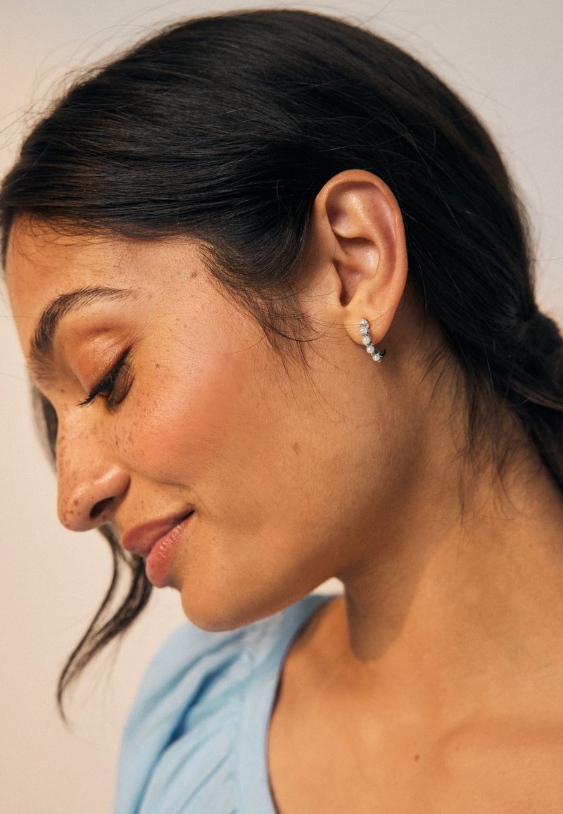 Silberner Creolen-Ohrring mit drei kleinen, runden, klaren Steinen, mit einem einfachen, geschwungenen Design, das sich sanft um das Ohrläppchen legt.