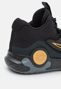 Sort atletiksko med en tekstureret mesh-overdel, mørkegrå detaljer, et gult rundt logo på sålen og et polstret ydersåledesign.