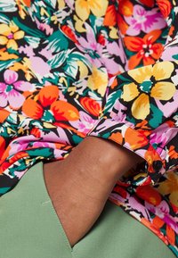 Mano di pelle scura in tasca di pantaloni verdi, indossando una camicia a maniche lunghe con vivaci motivi floreali in arancione, giallo, rosa e verde.