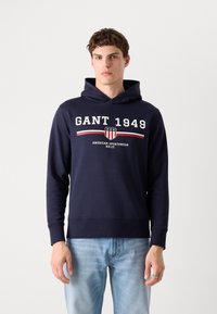 Sötétkék kapucnis pulóver fehér és piros "GANT 1949" felirattal. Puha anyagból készült, bordás mandzsettákkal és elöl kenguruzsebbel.