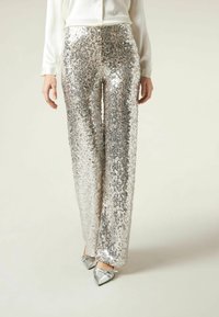 Kobieta w srebrnych spódnicach typu wide-leg ozdobionych cekinami, białej bluzce z długim rękawem i srebrnych szpilkach z ostro zakończonym noskiem stoi na tle jednolitą.