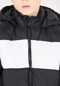 Veste matelassée noire et blanche avec capuche, dotée d'une fermeture éclair devant et d'un design en blocs de couleurs horizontaux. Texture lisse avec un look sportif.