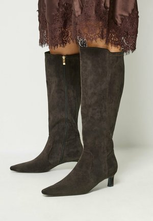 Femme portant des bottes hautes en daim marron foncé jusqu'aux genoux, avec des bouts pointus, petits talons et fermeture éclair latérale, associées à une jupe marron ornée de dentelle.