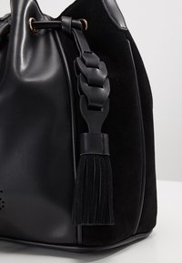 Musta käsilaukku, jossa on yhdistelmä sileää nahkaa ja pehmeää mokkaa, punottu olkahihna ja koristeellinen tupsu. Pyöreä muoto.