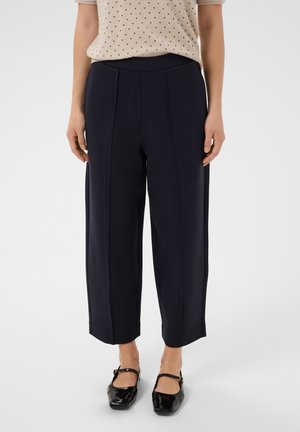 Femme portant un pantalon noir coupé, des chaussures Mary Jane noires, et une chemise beige à manches courtes avec des pois noirs.