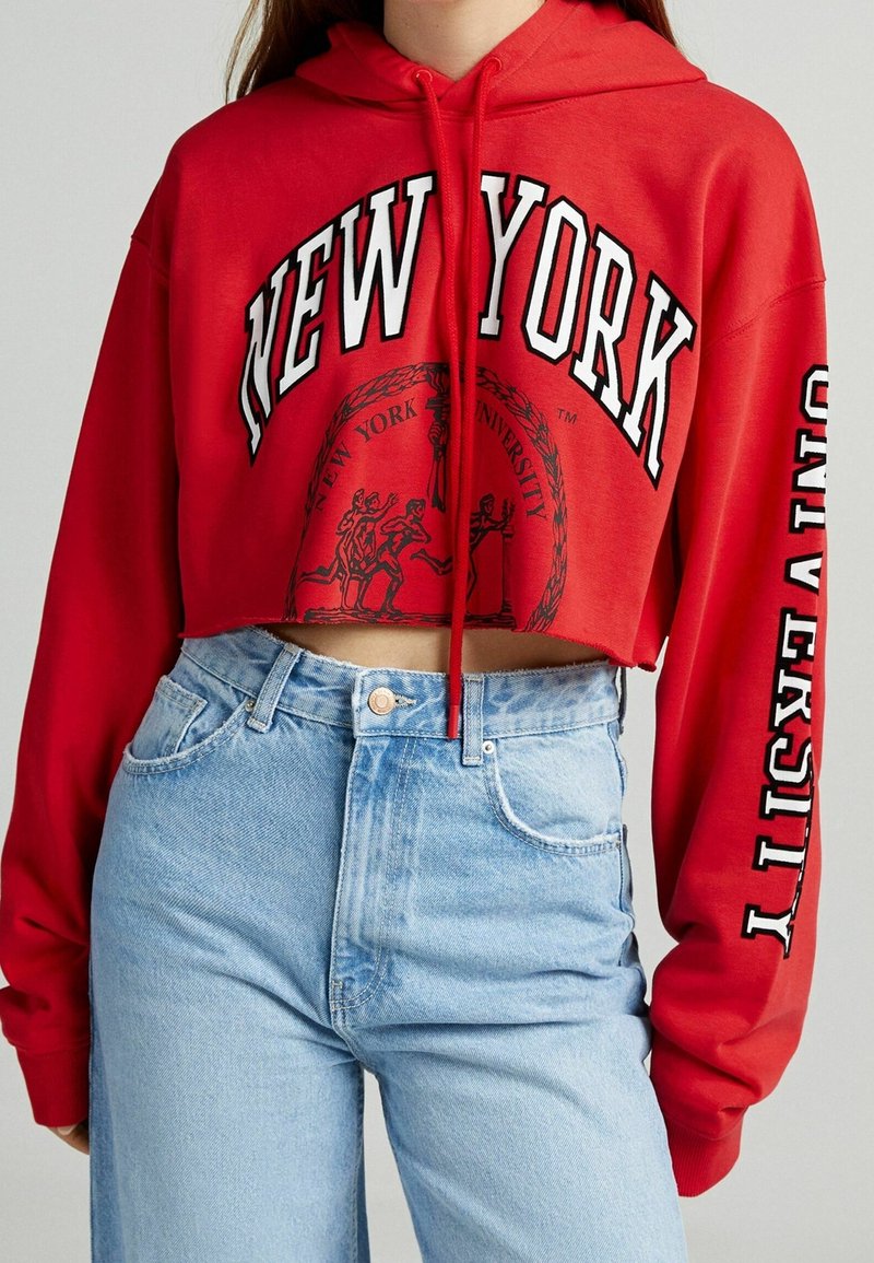 Personne portant un sweat à capuche court rouge avec le texte "New York University" et un jean taille haute bleu clair sur un fond uni.