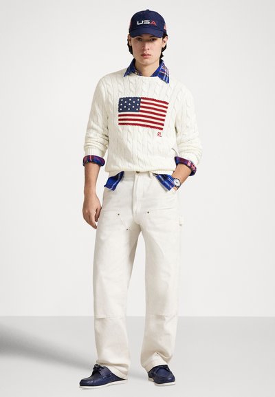 Camisola branca de malha com desenho da bandeira americana, usada sobre uma camisa azul aos quadrados, combinada com calças cor creme e sapatos azuis marinho.