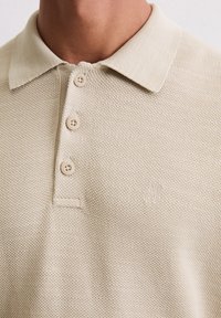 Beige Polohemd mit einem strukturierten Strickmuster, versehen mit einem klassischen Kragen, drei Knöpfen und einem kleinen Logo, das auf der linken Seite gestickt ist.