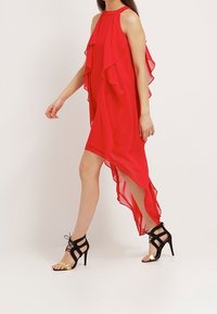 Robe en chiffon rouge avec des volants superposés et un ourlet asymétrique. Associée à des talons noirs à lanières avec des accents dorés. Texture lisse.