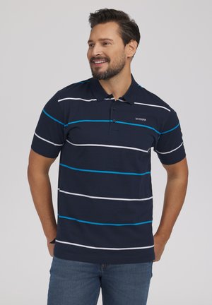 Mężczyzna uśmiechający się, ubrany w granatową koszulkę polo z białymi i niebieskimi poziomymi paskami oraz niebieskie dżinsy, ręce za plecami, neutralne tło.