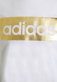 Camiseta blanca con una prominente banda rectangular metálica dorada con el logo de "adidas" en negrita y en color blanco. Textura de tela suave.
