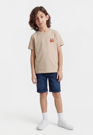 Chlapec so stredne dlhými hnedými vlasmi, oblečený v béžovom tričku s červeným motívom kraba, rifľových kraťasoch, bielych ponožkách a bielych teniskách, stojaci na bielom pozadí.