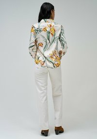 Blusa leggera color crema con motivo floreale, con fiori gialli e verdi, maniche lunghe e colletto. Abbinata a pantaloni crema e scarpe con tacco marroni.