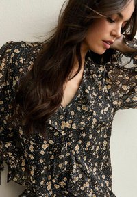 Czarna kwiatowa bluzka ozdobiona małymi żółtymi i białymi stokrotkami, z falbankami wzdłuż dekoltu i rękawów, wykonana z lekkiego, przezroczystego materiału.