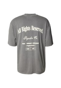 Šedé bavlněné tričko s velkým bílým nápisem na zádech. Design obsahuje nápis „Všechna práva vyhrazena“ a podrobnosti o značce tučným písmem.