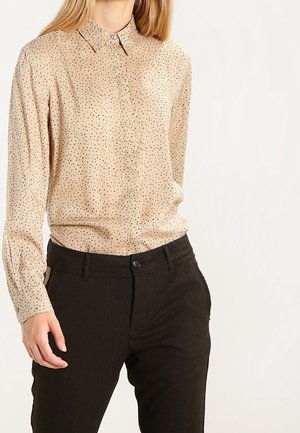 Personne portant une chemise beige à manches longues avec petits pois et un pantalon noir taille haute, debout avec une main relâchée.