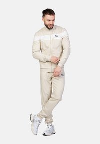 Completo sportivo beige realizzato in tessuto liscio, con una striscia bianca sul petto, composto da una giacca con zip e pantaloni coordinati con dettagli del logo.