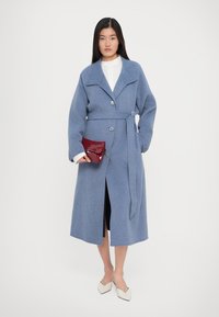 Femme portant un manteau en laine bleu ceinturé sur une blouse blanche, tenant une pochette rouge et des talons blancs à bout pointu, se tenant devant un fond uni.