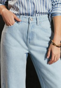 Jeans de denim de color azul claro, de talle alto, con pierna recta, bolsillos delanteros y cierre de botón. La textura está ligeramente desgastada.