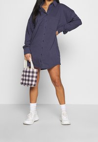 Kvinna som bär en oversized mörklila knappad skjortklänning, vita sneakers, vita strumpor och håller en liten pärlbeadad handväska med rutmönster.